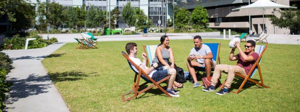 Four Indigenous students, three male and one female, sit on deck chairs on UTS Alumni Green, chatting and laughing. One is holding a football about to throw it back and forth with another male student. UTS buildings are in the background, including one showing the UTS sign.