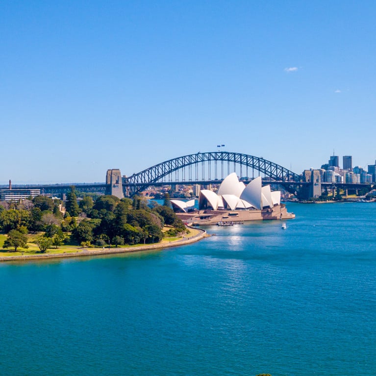 Panoramic view of Sydney's skyline featuring modern skyscrapers, the Sydney Opera House, and Harbour Bridge. A lush green park lines the waterfront under a clear blue sky.