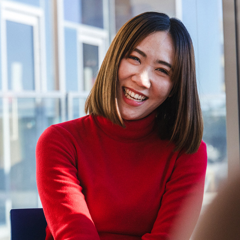 A woman in a red sweater smiles warmly while sitting indoors. A man nearby looks at his phone, laughing. Sunlight streams through large windows, creating a cheerful atmosphere.