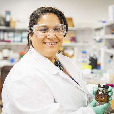 Photo of female researcher in safety goggles and lab coat, holding a labelled jar