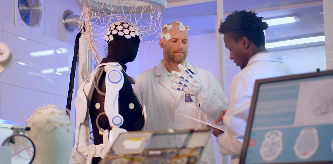 Two researchers in a lab one wearing a cap on head to test brain activity and wearing a glove. 