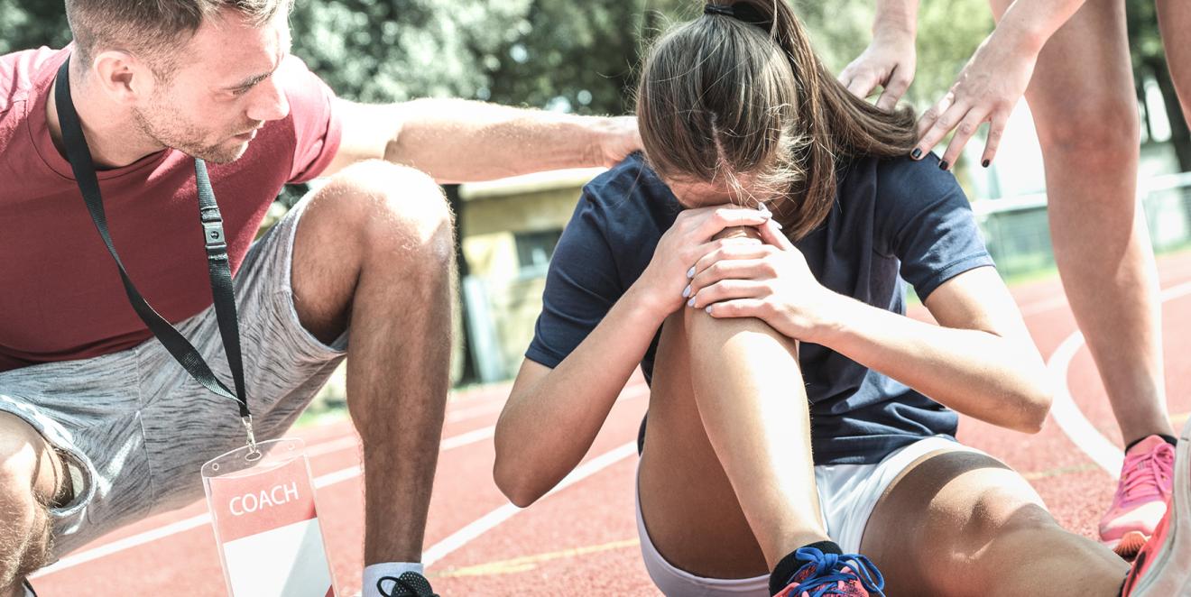 Injured runner sitting on a race track while two coaches comfort them.