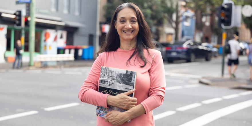 Woman standing at an intersection in a pink jumper holding a book and smiling and looking at the camera