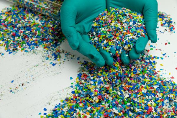 Two hands wearing green rubber gloves hold particles of coloured recycled plastic