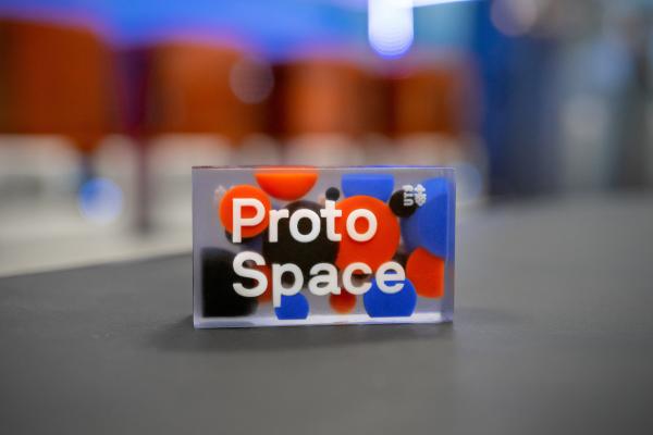 3D printed part, "ProtoSpace" in the ProtoSpace fab lab with 3d printers in background.