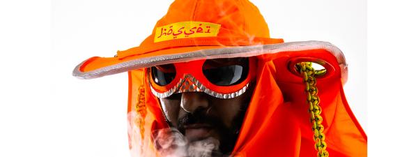 A figure wearing an orange hat and glasses blows smoke towards the camera. 