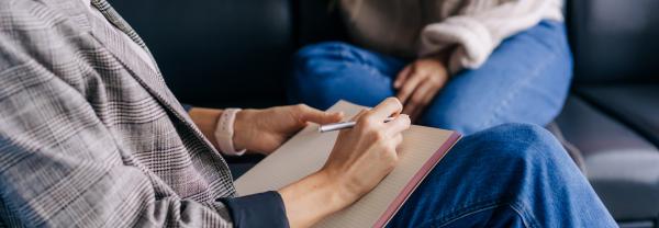 Close-up of someone's hands writing down notes while another person is sitting on a couch in the background. 