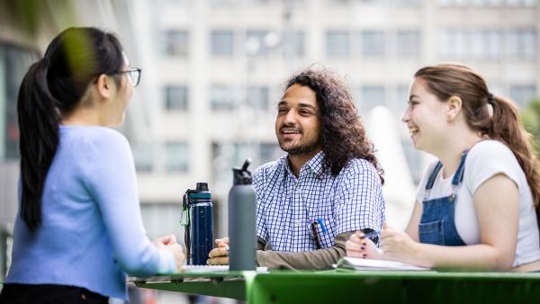 Three undergraduate students talk on tables near Alumni Green smiling and casual