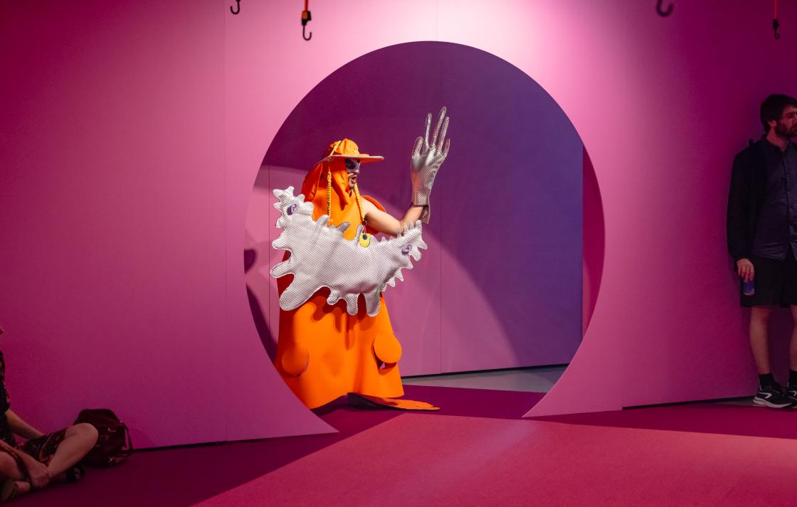 A person in an orange and silver costume emerges from a round doorway painted pink