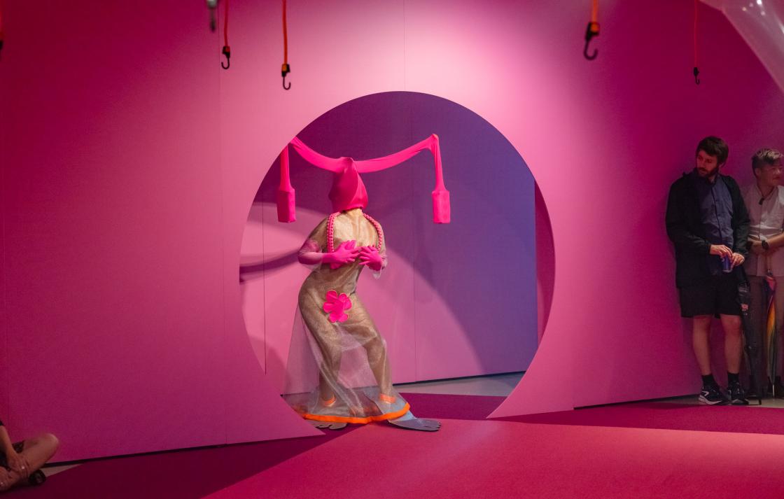 A person in a pink costume emerges from a round doorway painted pink