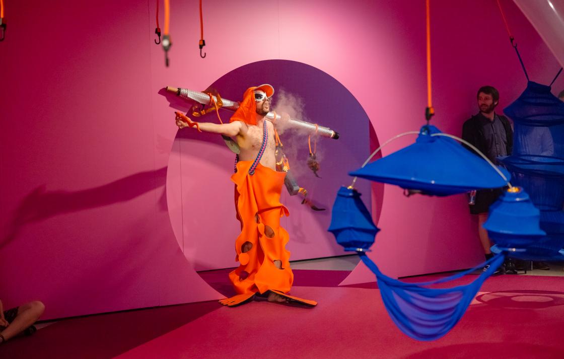 A person in an orange costume emerges from a round doorway blowing vape smoke