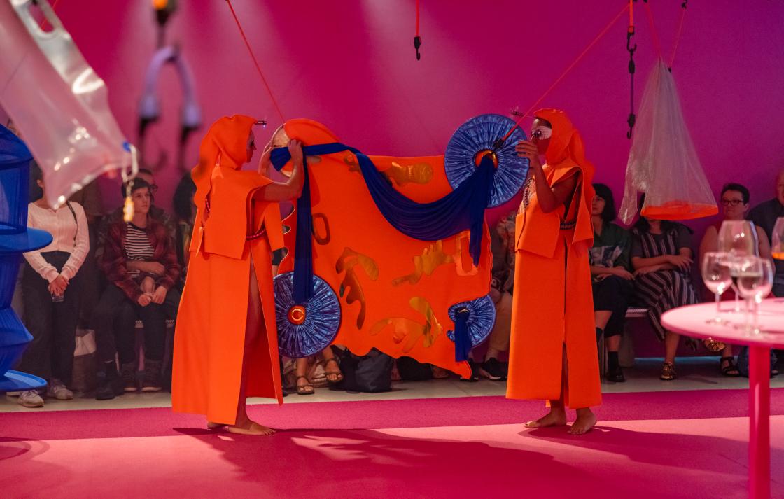 Two people in orange costumes hang another orange costume on octopus straps