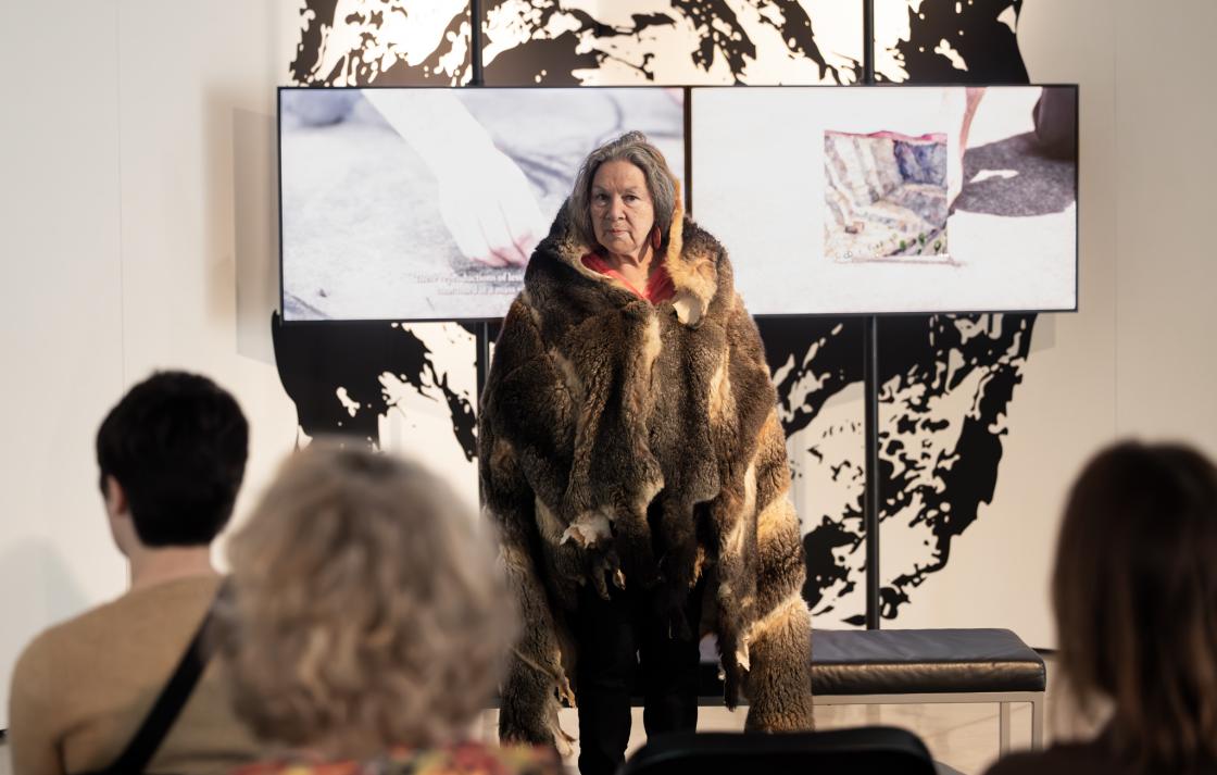 A woman in a possum skin cloak addresses and audience. Behind her are two screens. 