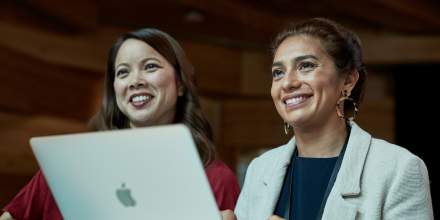 Two UTS female staff members smiling while looking off camera. One is typing on a Apple Mac laptop.