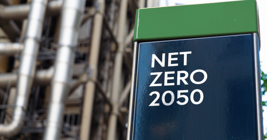 A green and blue shiny block reads "NET ZERO 2050" in white capital letters. In the background blurry pipes and infrastructure can be seen.