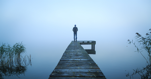 Man standing on the end of a jetting looking out over a foggy body of water. 