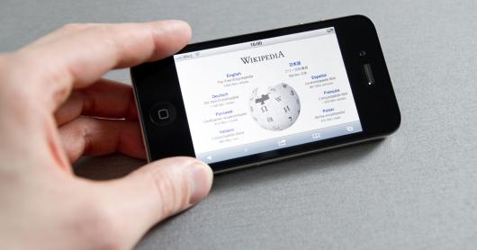 A person holds a mobile phone which displays the Wikipedia homepage.