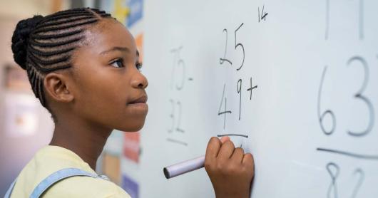 A young girl attempts a maths problem at a classroom whiteboard.