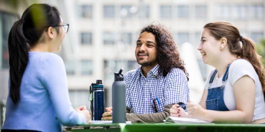 Three undergraduate students talk on tables near Alumni Green smiling and casual