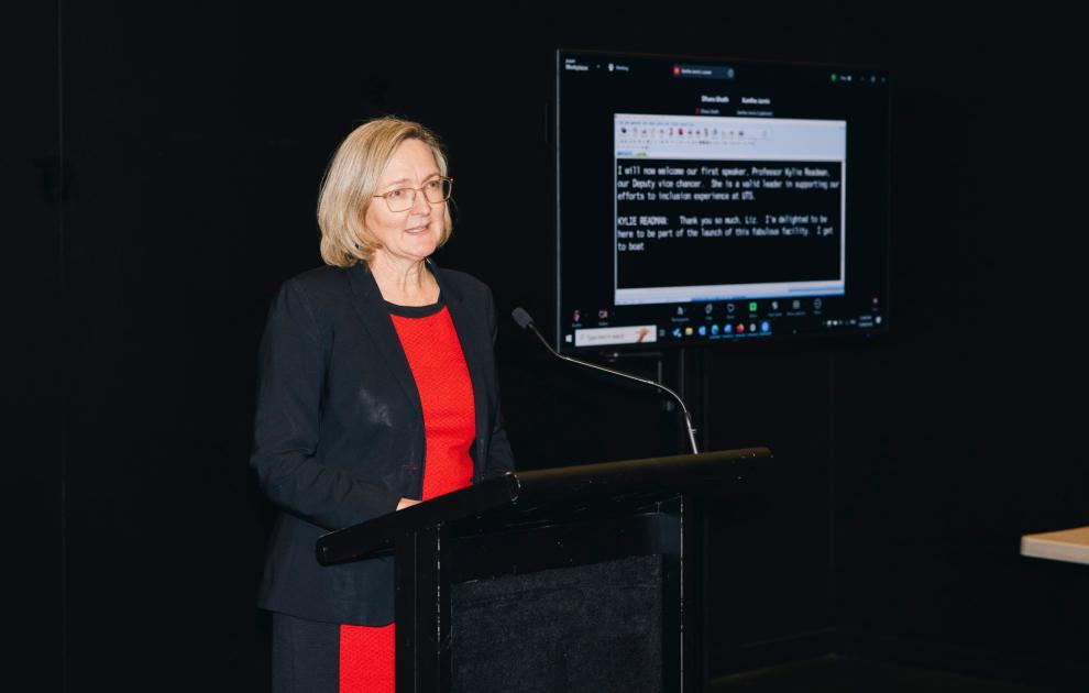 A woman with short blonde hair is speaking at a podium. She wears a red dress and a black blazer.