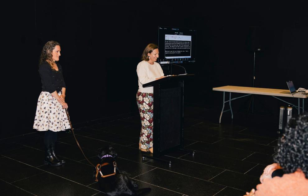 A woman holding the lead of a black guide dog is standing next to another woman speaking at a podium inside.