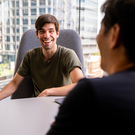 QSI's PhD student Fabio Henriques is sitting at a desk in conversation and smiling. In the background is a large window overlooking multistorey buildings opposite UTS Central on Broadway.