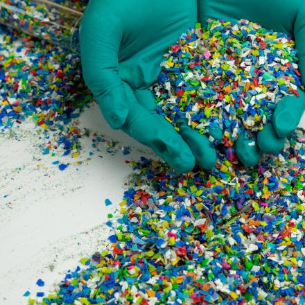 Two hands wearing green rubber gloves hold particles of coloured recycled plastic