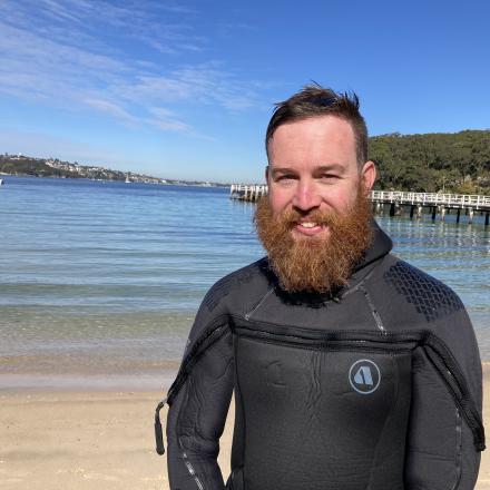 A man in a wetsuit stands on the beach with the ocean behind him