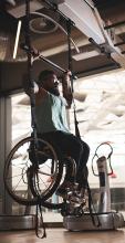 Man in a wheelchair uses a suspension training system to perform pull-ups in a gym.