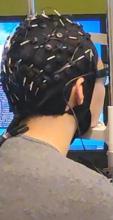 A person equipped with an EEG cap is positioned in front of two computer screens, possibly monitoring brainwave data.