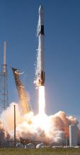 A space rocket launches from the ground, soaring upward against a backdrop of blue sky and billowing smoke.