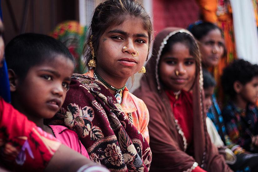 Children in traditional Indian dress sit outside a home, looking directly at camera