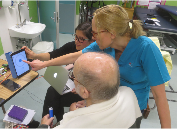 Female physiotherapist and female student point to digital screen, showing an elderly male patient the results of his stroke rehabilitation.