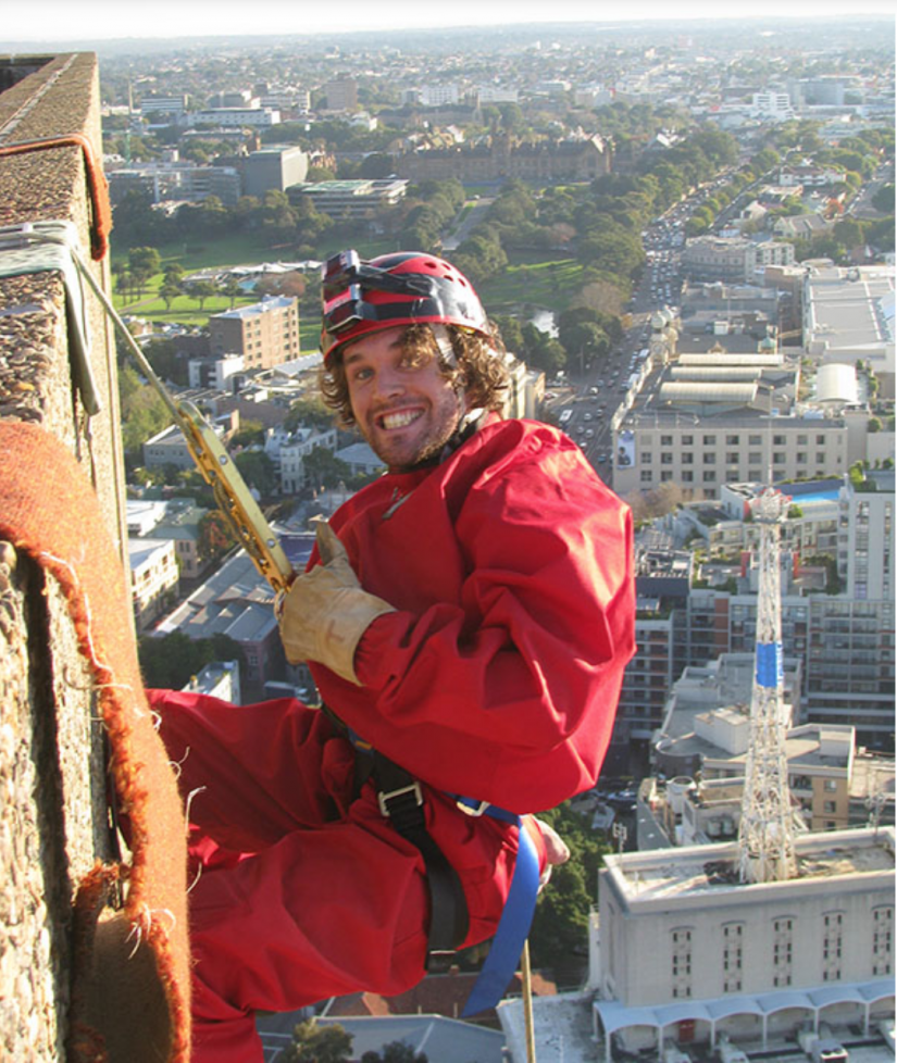 Young man dressed in red, hanging off the top of the UTS Tower, smiling at camera