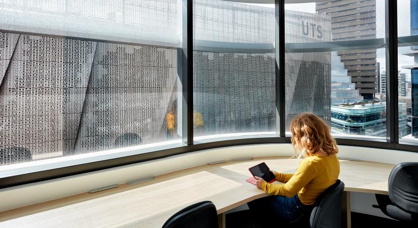 A woman with a digital tablet works at a desk bathed in sunshine, with large windows looking out to UTS Buildings 11, 2 and 1