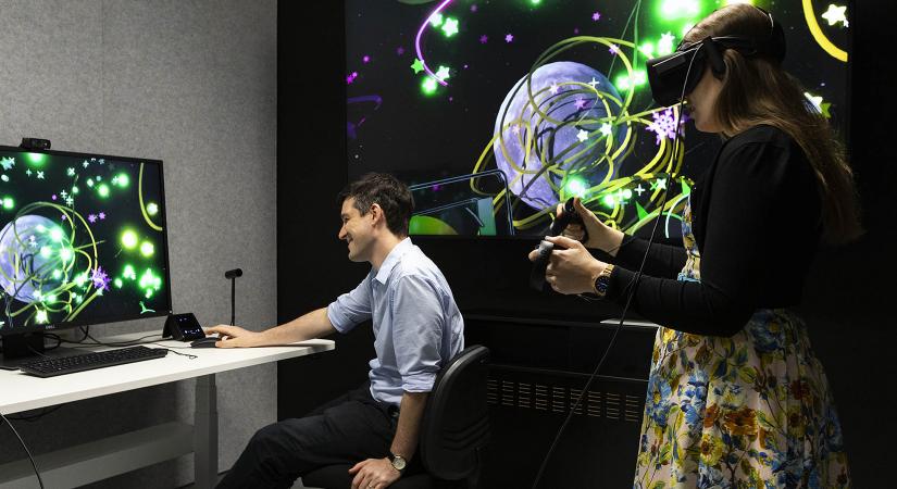 A young woman wears a virtual reality headset and holds two hand controllers, while a man operates a computer with a digital screen showing chemical structures