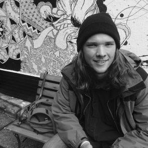 Image of Taylor Thompson-Fuller sitting in on a park bench against a wall with street art. Image is in black and white.