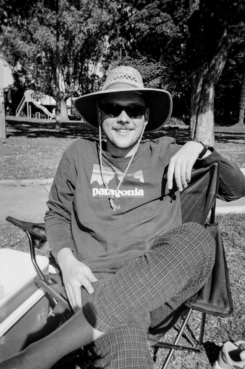 Taylor Thompson-Fuller, FASS graduate - reclining in a chair outside and wearing a hat and sunglasses - image is in black and white