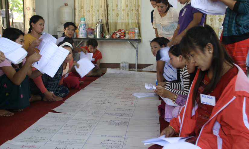 A group of Thai women sit on the floor, writing on a large piece of paper