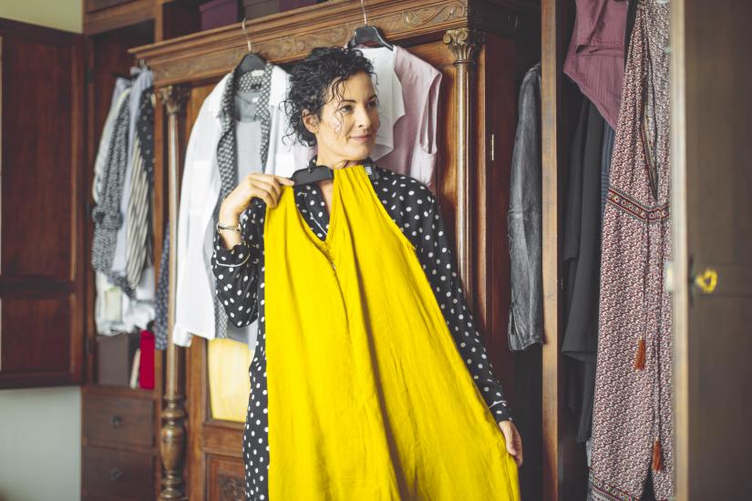 A woman holds up a yellow dress. There is an open wardrobe behind her.