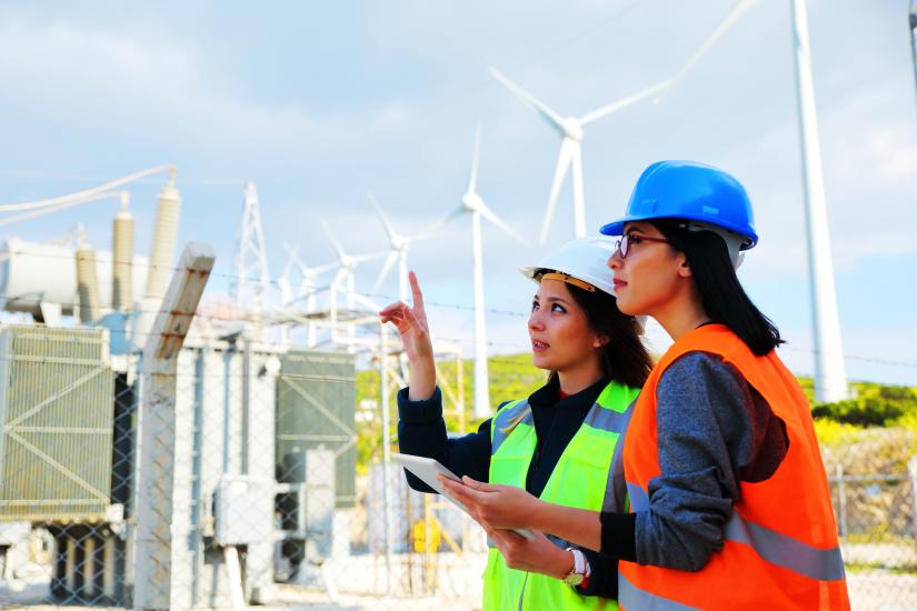 Two female energy workers in hi-vis jackets and hard hats stand in front of wind turbines