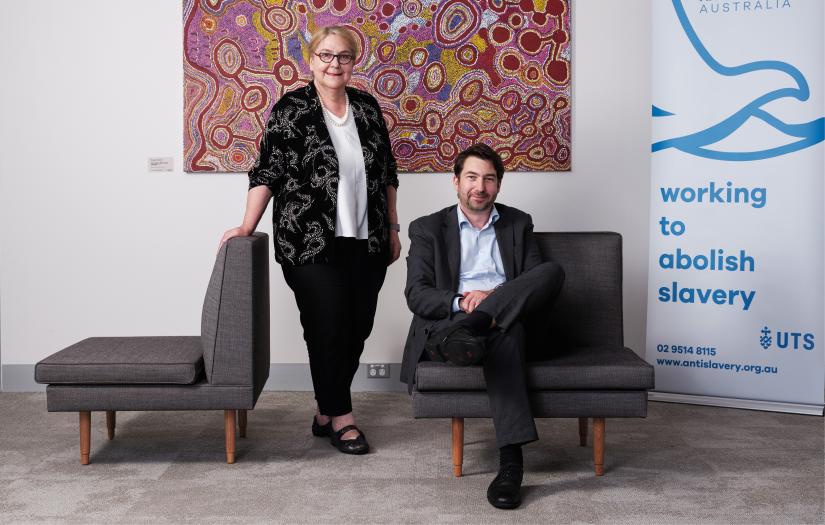 Woman standing leaning on a grey armchair standing next to a seated man in front of colourful indigenous artwork and a pull up banner