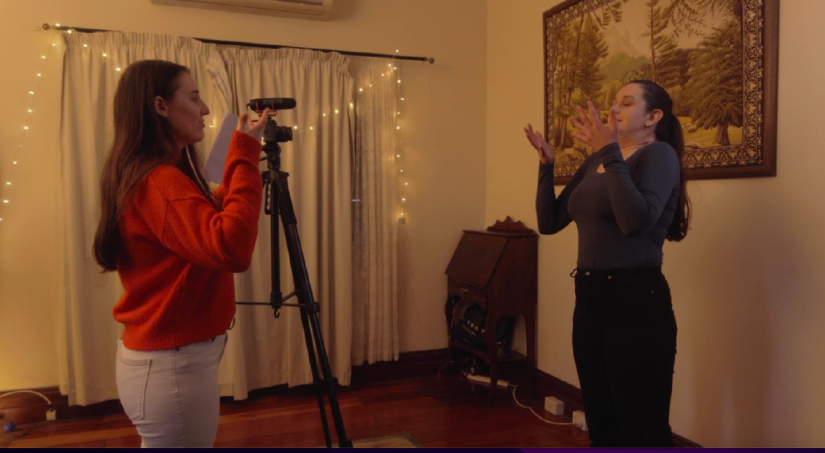 One woman with a video camera shooting footage of another woman. Woman behind camera wears a red jumper. Woman on camera in purple jumper and is raising her arms and closing her eyes. They are inside a lounge room.