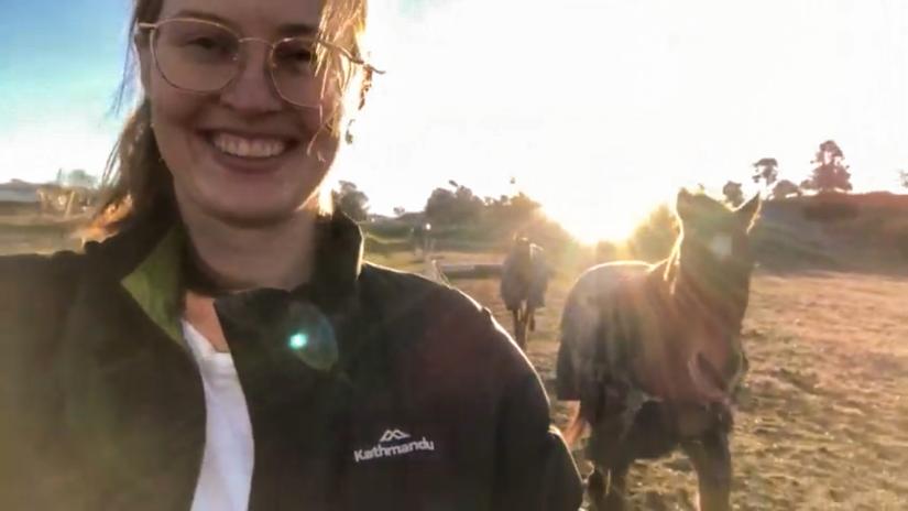 A young woman smiles at the camera, the sun and horses in the background.