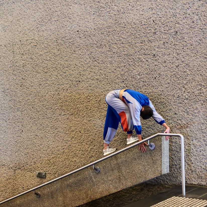 A dancer in a grey and blue tracksuit leans against a stair rail