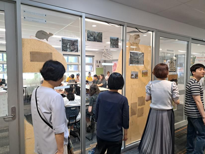 image of four people walking along glass walls and on the other side are students in a classroom