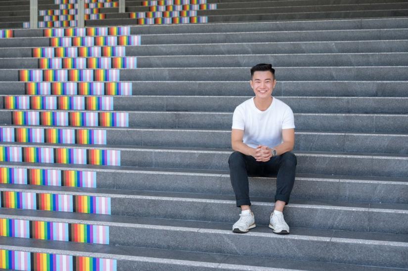 Photo of young male sitting on grey steps. There is a painted rainbow on the steps next to him.