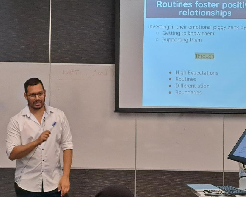 image of a man giving a lecture at the front of a classroom. The screen behind him displays a slide with information about using routines to foster positive relationships.