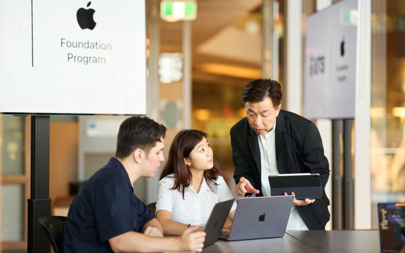 Two students looking at Apple laptops while a course instructor points to the screen.