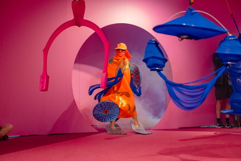 Figure dressed in an orange cloak, yellow cap with flaps that cover the face. Figure wearing sunglasses, big silver flippers and holding shiny blue shield. Against a pink red room, with pink, blue contraptions hanging from the ceiling.